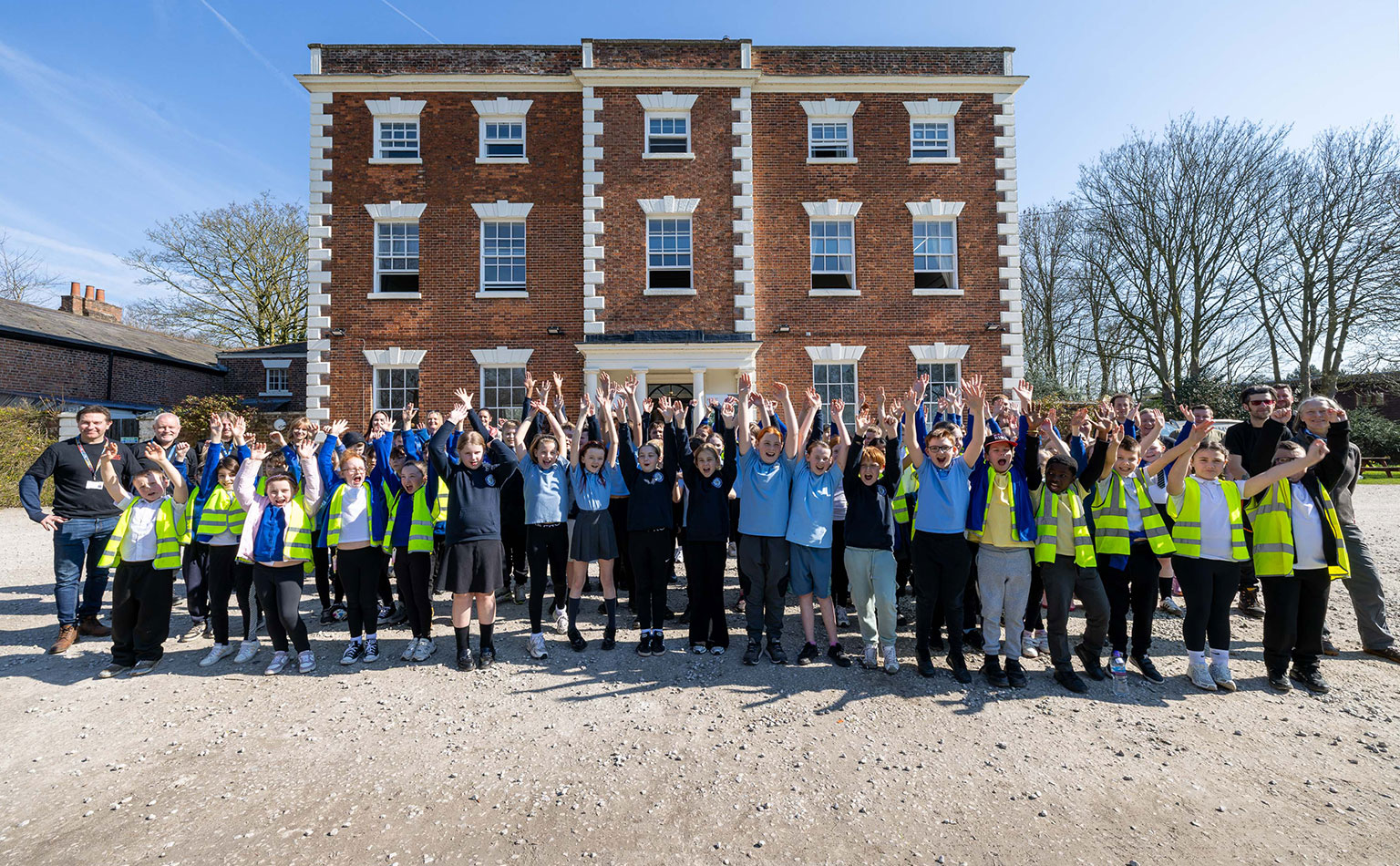 A large group of school children, cheering, with their hands in the air whilst standing in front of Trafford Hall.
